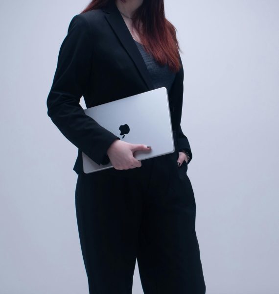Businesswoman in a dark suit holding a laptop against a neutral background, symbolizing modern work culture.